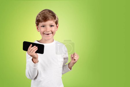 Foto de Retrato de niño feliz con el puño levantado y el teléfono en la mano, copia el espacio de fondo verde vacío. Concepto de educación y éxito - Imagen libre de derechos