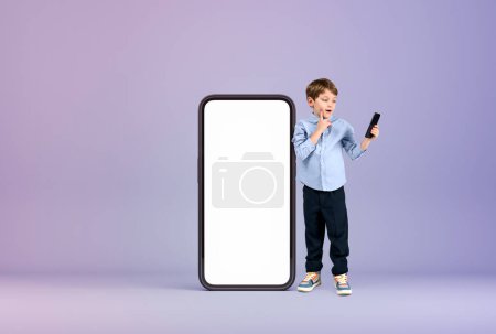 Photo for School boy with pensive look, standing near big smartphone mock up copy space screen, lilac background. Concept of mobile app and e-learning - Royalty Free Image