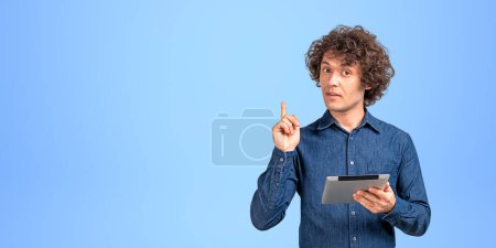 Téléchargez les photos : Jeune homme intéressé tenant tablette à la main, doigt pointant vers le haut recommander produit ou service en ligne. Copier l'espace fond bleu. Concept d'offre et de conseil - en image libre de droit