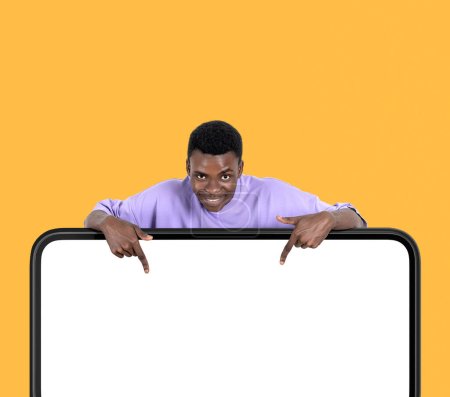 Téléchargez les photos : Homme noir américain souriant, doigt pointant vers le bas à grand écran numérique blanc maquette sur fond jaune. Concept d'abonnement et de suivi - en image libre de droit