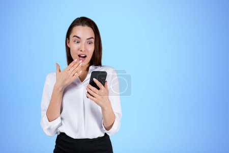 Téléchargez les photos : Femme d'affaires confus portant des vêtements formels est debout toucher menton tenant smartphone près du mur bleu vide en arrière-plan. Concept de modèle surpris, homme d'affaires étonné, femme choquée - en image libre de droit