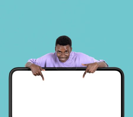 Foto de Hombre afroamericano sonriendo, el dedo apuntando hacia abajo en gran simulacro de pantalla digital espacio de copia, fondo turquesa. Concepto de conexión en línea y red - Imagen libre de derechos