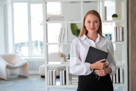 Photo for Young businesswoman student holding notebook in hands, smiling portrait looking at the camera. Minimalist office room on background. Concept of business education - Royalty Free Image