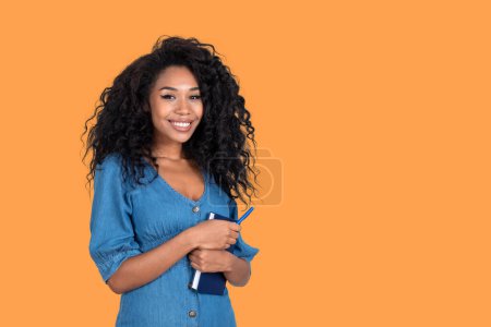 Photo for Happy blank woman student with textbook and pen in arms, smiling portrait looking at the camera. Empty copy space orange background. Concept of business student and career - Royalty Free Image