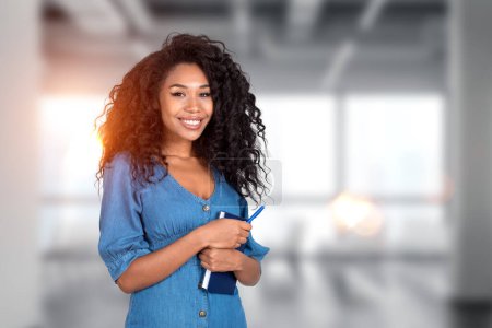 Téléchargez les photos : Femme d'affaires noire tenant un manuel et un stylo, souriant portrait étudiant sur fond de bureau flou. Concept de formation et d'études commerciales - en image libre de droit