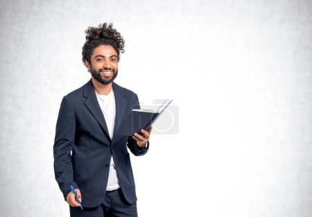 Photo for Smiling arab businessman with notebook and pen in hands, looking at the camera on empty grey concrete wall background. Concept of business education - Royalty Free Image