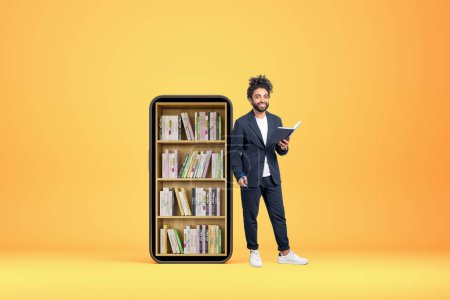 Photo for Middle eastern businessman full length standing with notebook and pen in hands, yellow background. Large smartphone screen with books on shelf. Concept of e-learning - Royalty Free Image