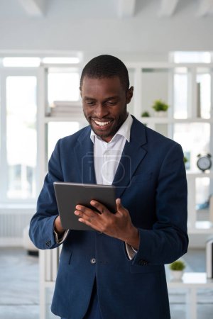 Téléchargez les photos : Un homme d'affaires afro-américain souriant portant un costume officiel est debout tenant et tapant sur une tablette au bureau. Concept de modèle, homme d'affaires, gestion du temps, appareil, gadget, patron réussi - en image libre de droit