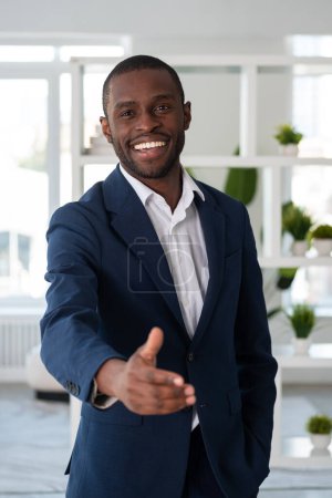 Téléchargez les photos : Un homme d'affaires afro-américain souriant portant un costume officiel se tient debout la main tendue pour une poignée de main sur le lieu de travail de bureau en arrière-plan. Concept de modèle, homme d'affaires, réalisateur, patron, salutation - en image libre de droit