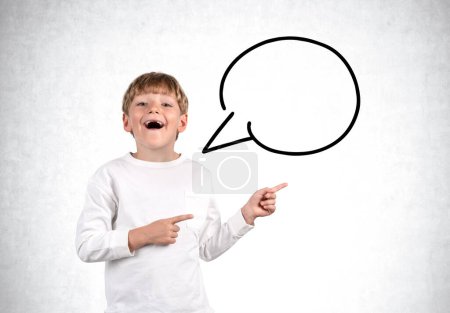 Photo for Smiling school boy with opened mouth, looking at the camera and fingers pointing to the side. Copy space speech bubble on grey concrete wall background. Concept of feedback - Royalty Free Image