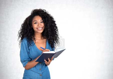 Téléchargez les photos : Heureuse femme noire prenant des notes, souriant portrait étudiant d'affaires en regardant la caméra. Espace de copie fond de mur en béton gris vide. Concept d'études et de cours - en image libre de droit