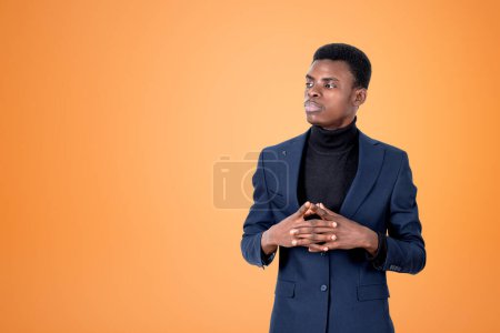 Foto de Reflexionar empresario afroamericano con ropa formal de pie con las manos en la pose de bloqueo cerca de la pared naranja vacía en el fondo. Concepto de modelo, persona de negocios exitosa, pensando en los negocios - Imagen libre de derechos