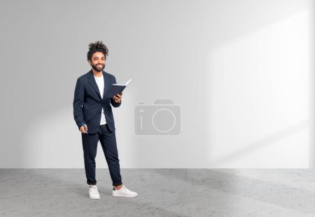 Photo for Middle eastern businessman full length standing on grey concrete floor. Notebook and pen in hands, smiling looking at the camera on copy space white background. Concept of business startup - Royalty Free Image