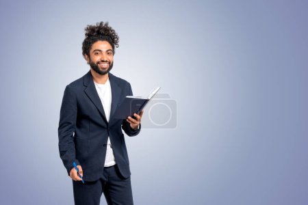 Foto de Hombre de negocios de Oriente Medio con cuaderno y pluma en las manos, sonriendo mirando a la cámara en el espacio de copia vacío fondo azul. Concepto de educación y carrera. - Imagen libre de derechos