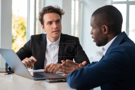 Photo for Serious African American businessman wearing formal suit talking to colleague explaining on laptop at office workplace with documents and smartphone in background. Concept of teamwork, cooperation - Royalty Free Image