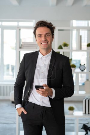 Téléchargez les photos : Un homme d'affaires souriant portant un costume formel tient son smartphone en regardant la caméra sur le lieu de travail du bureau. Pose main dans la poche. Concept de modèle, homme d'affaires, gestion du temps, appareil, gadget - en image libre de droit
