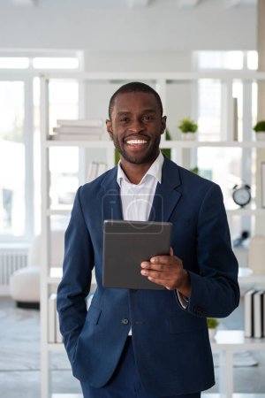 Téléchargez les photos : Souriant homme d'affaires afro-américain portant un costume formel est debout tenant appareil tablette regardant la caméra sur le lieu de travail de bureau. Concept de modèle, homme d'affaires, gestion du temps, appareil, gadget - en image libre de droit