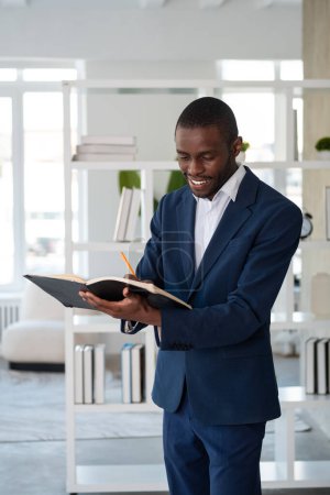 Photo for Smiling African American businessman wearing formal suit is standing taking notes in notebook at office workplace in background. Concept of model, business person, time management, boss, director - Royalty Free Image