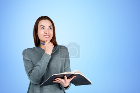 Photo for Thoughtful and smiling woman looking up with notebook in hands and hand to chin, copy space empty blue background. Concept of thoughts and idea - Royalty Free Image