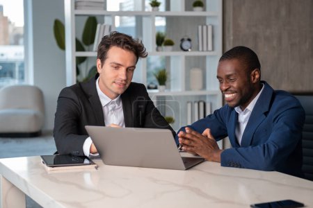 Téléchargez les photos : Homme d'affaires afro-américain souriant en costume officiel parlant à un collègue expliquant le projet sur ordinateur portable au bureau avec un appareil tablette en arrière-plan. Concept de travail d'équipe, coopération, coworking - en image libre de droit
