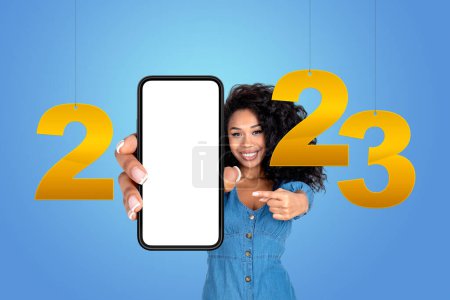Téléchargez les photos : Jeune femme noire doigt pointant vers le smartphone blanc maquette, numéros dorés 2023 année suspendus sur fond bleu. Concept de vacances et offre d'affaires - en image libre de droit