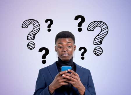 Téléchargez les photos : Jeune homme d'affaires noir portrait avec smartphone, étonné et confus. Jeu de points d'interrogation sur fond violet. Concept de problème et de solution - en image libre de droit