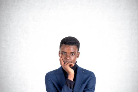 Foto de Retrato de un hombre de negocios afroamericano joven y reflexivo que piensa de pie con la mano en la barbilla de pie sobre el fondo de la pared de hormigón. Concepto de planificación y contemplación - Imagen libre de derechos