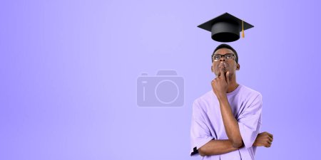 Foto de Retrato de un joven estudiante universitario afroamericano pensativo con gafas paradas sobre el fondo del espacio de copia púrpura y mirando el mortero. Concepto de educación, graduación y elección de carrera - Imagen libre de derechos