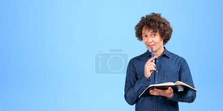 Photo for Thoughtful businessman with notebook and pen on chin, curious looking at the camera on copy space blue background. Concept of plan, idea and management - Royalty Free Image