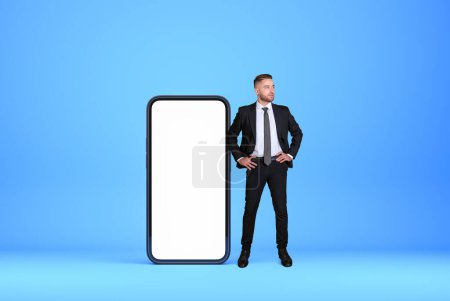 Photo for Confident handsome businessman wearing formal wear standing in hands on hips pose near smartphone with mockup and empty blue wall in background. Presentation of mobile application, social media - Royalty Free Image