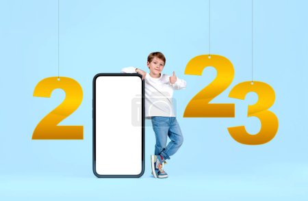 Photo for Child boy smiling with a thumb up, 2023 numbers hanging on blue background. Smartphone display with mock up copy space screen. Concept of new year and mobile app - Royalty Free Image
