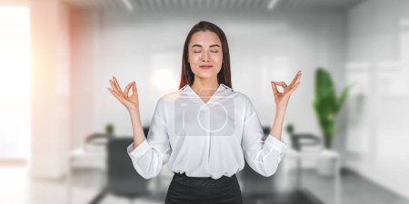 Foto de Mujer de negocios meditando en la sala de oficina en el fondo. Mujer sonriente con los ojos cerrados y dedos de loto. Concepto de relax después del trabajo, yoga y autocontrol - Imagen libre de derechos