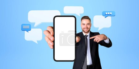 Téléchargez les photos : Portrait de joyeux jeune homme d'affaires pointant vers le smartphone avec écran maquette avec des bulles de parole au-dessus de lui debout sur fond bleu. Concept de médias sociaux et de bavardage - en image libre de droit