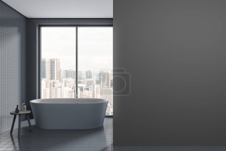 Téléchargez les photos : Intérieur de la salle de bain élégante avec des murs en carrelage gris et le sol, baignoire blanche confortable debout près de la grande fenêtre et copier mur d'espace sur la droite. Rendu 3d - en image libre de droit