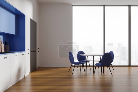 Téléchargez les photos : Intérieur de cuisine moderne avec murs blancs, parquet, armoires blanches confortables, placards bleus, grand réfrigérateur et table à manger ronde avec chaises bleues debout près de la fenêtre panoramique. Rendu 3d - en image libre de droit