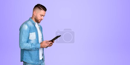 Photo for Young man student using tablet, handsome guy in denim shirt on empty copy space purple background. Concept of internet, communication and online education - Royalty Free Image