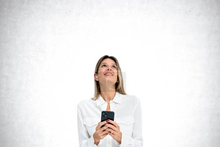 Téléchargez les photos : Portrait d'une jeune femme d'affaires européenne heureuse avec smartphone et écouteurs debout sur un fond de béton et regardant vers le haut. Concept de communication et médias sociaux - en image libre de droit