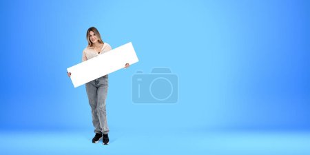 Photo for Young smiling woman standing full length, holding a mockup blank signboard in hands on empty blue background. Concept of recommendation, announcement and sign - Royalty Free Image