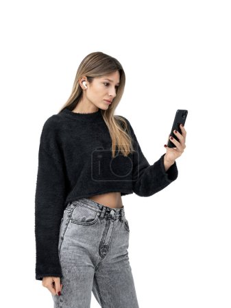 Photo for Serious woman looking at smartphone in hand, side portrait wearing earbuds. Isolated over white background. Concept of online communication, mobile conference and video call - Royalty Free Image