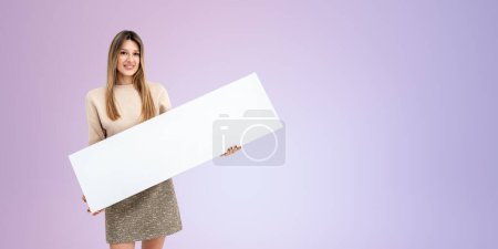 Téléchargez les photos : Heureuse femme souriante tenant une bannière de panneau blanc maquette dans les mains, espace de copie vide fond violet. Concept de vente, publicité, annonce et recommandation - en image libre de droit