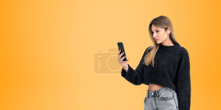 Photo for Portrait of serious young European woman in casual clothes making video call with smartphone and headphones over yellow copy space background. Concept of communication and social media - Royalty Free Image