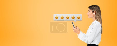 Photo for Side portrait of beautiful young and smiling European businesswoman holding smartphone with five star rating above it over yellow copy space background. Concept of product and service evaluation - Royalty Free Image
