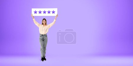 Foto de Retrato de cuerpo entero de una joven europea feliz vestida con ropa casual con cinco estrellas encima de su cabeza sobre el fondo del espacio de copia púrpura. Concepto de evaluación de productos y servicios - Imagen libre de derechos