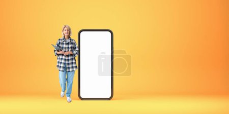 Photo for Smiling woman teacher or student standing near large mock up smartphone, blank empty screen on orange background. Concept of education, online learning and mobile app - Royalty Free Image