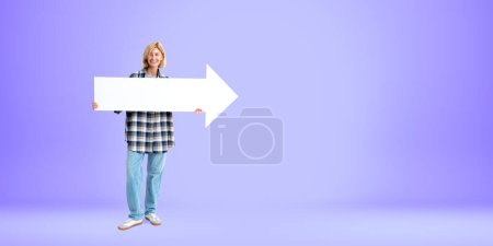 Foto de Mujer rubia sonriente de pie de cuerpo entero, sosteniendo un letrero de flecha en blanco maqueta en las manos sobre un fondo púrpura vacío. Concepto de dirección, punto y recomendación - Imagen libre de derechos