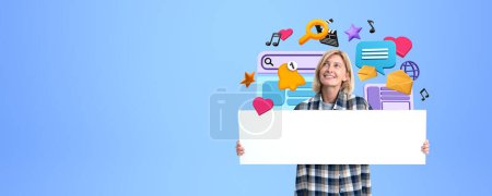 Photo for Happy young woman showing mockup blank signboard, looking up at colorful cartoon social media and internet icons on copy space blue background. Concept of communication and message - Royalty Free Image