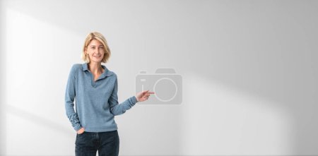 Téléchargez les photos : Portrait de heureuse jeune femme européenne pointant sur le côté debout près du mur blanc de l'espace de copie. Concept de publicité sur les produits et services - en image libre de droit