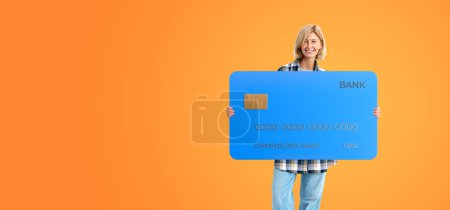 Photo for Happy smiling blonde woman holding a blue bank credit card, copy space empty orange background. Concept of payment, purchase, finance and electronic money - Royalty Free Image