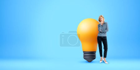 Foto de Retrato de una joven pensativa de pie cerca de una gran bombilla sobre el fondo azul del espacio de copia. Concepto de idea brillante, planificación y lluvia de ideas - Imagen libre de derechos