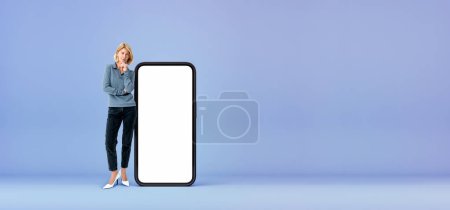 Foto de Mujer europea joven reflexiva de pie cerca de un gran teléfono inteligente con pantalla simulada sobre el fondo del espacio de copia púrpura. Concepto de comunicación y anuncio de sitios web y aplicaciones - Imagen libre de derechos